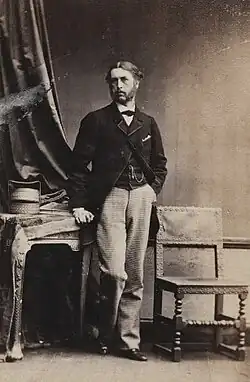 Standing photographic studio portrait, wearing a jacket and waistcoat, between a table and a chair