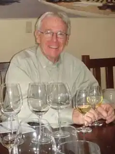 A man with white hair and glasses wearing a brown button-down shirt, smiling at the camera