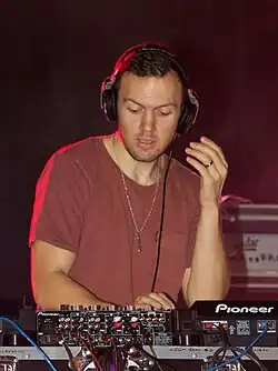 Chris Lake, a white man, stands in front of a soundboard, wearing a red t-shirt and headphones.