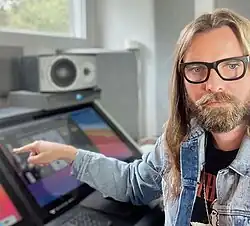 Chris Traynor wearing a black print t-shirt and denim jacket, looking at camera with set face and pointing sideways at a computer screen with right arm extended.