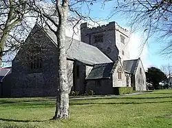 A low cruciform pale-coloured church seen from the southeast with the chancel prominent, beyond which is a broad castellated tower; in the foreground is the trunk of a tree standing in a grassed area