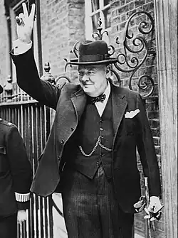A jowly, well-dressed man, obviously Winston Churchill, standing outside a doorway. He is smiling and making a "V for victory" gesture.