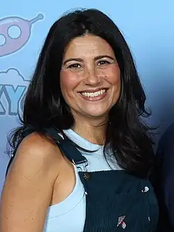 A dark-haired white woman wearing a white tank top and blue overalls smiling towards the camera.