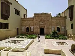 Exterior view of multi-story stone building with Islamic architectural features, showing courtyard area with plantings in foreground