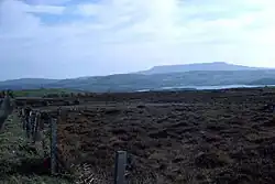 Photograph of a long, flat mountain ridge on the far horizon, with layers to the fore: foothills of the aforementioned mountain; a sliver of a lake; blanket bog and a fence in the very near foreground.