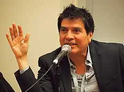 A middle-aged Chicano man with tousled hair speaks into a microphone. He has his right elbow resting on the table in front of him, his right palm facing out towards the audience.