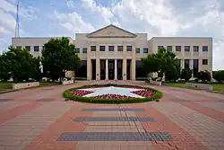 Fronts picture of a two-story administration building on a cloudy day. The walkway is shown leading up to the building including a circular garden with white flowers forming a star.