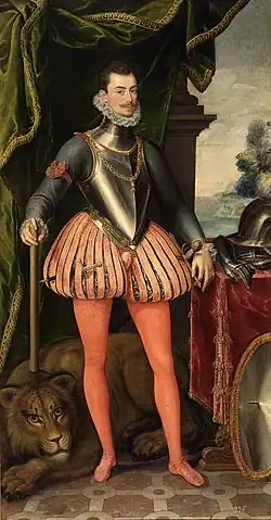 A tall brown-haired man in armour and a royal attire posing for a painting. He is standing under a luxurious green drapery and wearing a red brassard on his right arm. An attentive lion is lying behind him at his feet. His helmet and gloves are lying on a table beside him while his shield is resting on the floor in front of the table.