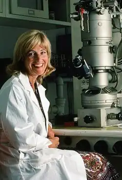 A smiling blonde older woman in a lab coat sits at an electron microscope.