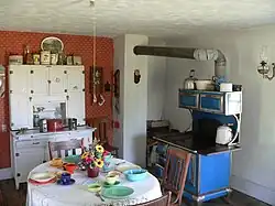 Interior of kitchen: cupboard against wall, table and chairs in foreground, cast-iron stove with pipe leading to chimney