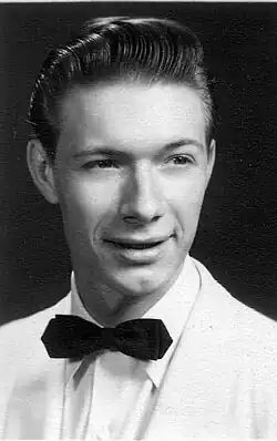 He is smiling with his mouth slightly open, and his left eyebrow is slightly raised. His hair is gelled back, and he wears a white suit with a black bowtie.