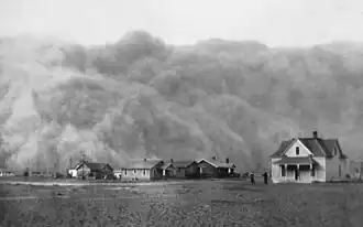 A dust storm approaches a collection of houses, dwarfing them in height.