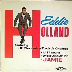 Holland seated on a logo of his name, smiling and wearing a suit