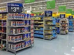 Aisles in a supermarket with small sets of shelves on the end, receding from left to right. Atop each set of shelves is a large white on black sign with the price of an item for sale on those shelves. White on green signs with the types of products in the aisles hang from the high ceiling.