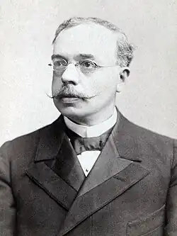A black and white image of a man in his late middle age. He has formal dress small glasses and a moustache.