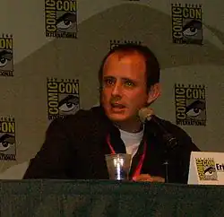 A man sitting in front of a microphone. His hands are folded and he is speaking.