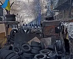 Photograph of security patrols standing behind a makeshift barricade of tires, burned vehicles, cardboard, and wooden pallets.