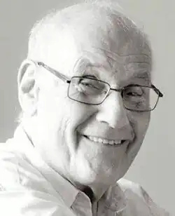Three-quarter profile, black and white, publicity headshot of a clean-shaven, elderly, bespectacled, smiling, bald man.