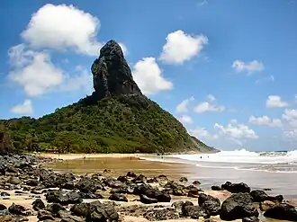 In the foreground, a wavy sea at the right and a yellow beach covered with black stones at the left. In the background, a mountain covered with green vegetation out of which towers a steep black rock.