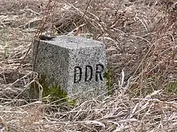 Square grey stone with "DDR" carved on one face, located in a patch of scrubby brown grass