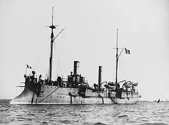 A large ship with two tall, thin smoke stacks and two light masts sits at anchor. It has several small boats hoisted on derricks toward the rear of the ship.