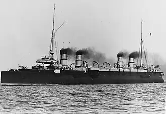 A large, black ship with two pairs of widely spaced funnels belching thick black smoke cruises through calm water.