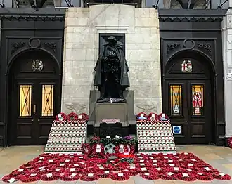 Bronze statue against a white stone wall with oak panels in the background and poppy wreaths on the floor below