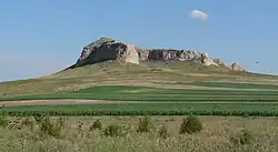 Steep-sided rocky bluff surrounded by grassland; green fields in foreground
