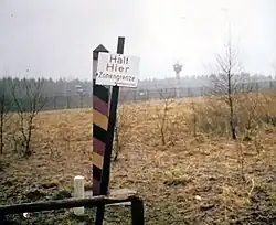 View across a landscape with a leaning sign reading "Halt Hier Zonengrenze Bundesgrenzschutz" in the foreground, a red/black/yellow striped square-shaped pole just behind, and a metal fence and watchtower visible across a strip of open ground in the background