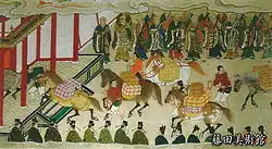 Horses laden with packages being led towards a gate. Their path is lined with monks.