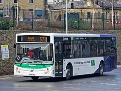 White and blue single deck bus with angular treen striping on front and sides passing through a bus station.