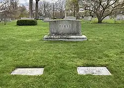 A photograph of a green cemetery sward with two flat gray headstones side by side. The left headstone bears the inscription "John T. Pirie Jr.", "May 22, 1903" - "Nov 10, 1980", and the right headstone bears the inscription "Ginevra King Pirie", "November 30, 1898" - "December 13, 1980". Behind them looms a larger upright headstone inscribed with the single word "Pirie".