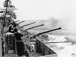 The flank of a ship. Several long-barrelled guns are aimed over the side, and are being operated by sailors. One of the guns has just fired, with a cloud of smoke issuing from the barrel.