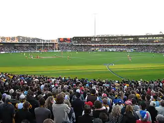 A small stand to the left and a two tier stand and scoreboard filled with people in the backdrop of an oval grass playing surface scattered with players. Spectators stand in the foreground.
