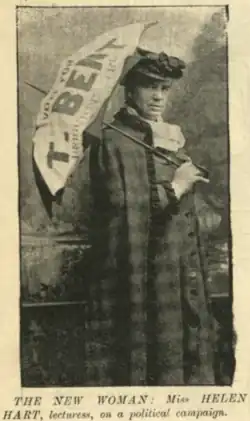 grainy image from a newspaper of a woman standing, facing to her left, but looking at the camera. She is expressionless, and wearing a dark coat and cloak with a white ruff collar. She is holding a large umbrella over her shoulder. The umbrella says "Vote for T. Bent" and further writing is obscured.