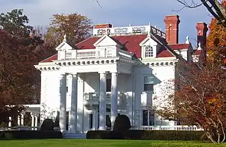 A white house with a colonnaded front and dormer windows in its red roof with brick chimneys on top seen from slightly to the right of center. The sun lights the house from the left and on either side are trees showing fall color