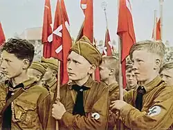 Young boys, wearing brown Nazi uniforms, on parade with red swastika banners