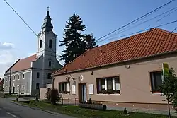 Tannish square one-story building with red shingles. A white Lutheran church with a steeple is at left.