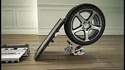 a flat board strikes the tread of a car tyre at a 45-degree angle. The tyre is balanced on top of a smaller piece of machinery on a wooden floor.