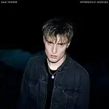 A Caucasian man in a black shirt and a denim jacket is shown, amidst a black background. "Sam Fender" and "Hypersonic Missiles" are displayed in white text in the top left and top right corners, respectively.