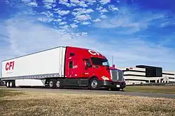 Tractor-trailer in front of large, multi-story office building, CFI headquarters in Joplin. Tractor is painted red with a white CFI logo on the face of the roof fairing. Trailer is painted white with a red CFI logo on the side near the tail.