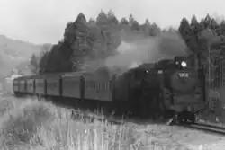 A monochrome photograph of a large 4-6-4 steam locomotive hauling seven coaches along a single track alongside a forest.