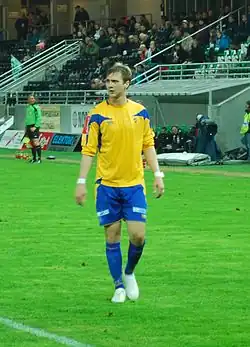 A brown-haired Caucasian American is standing in the center of a grass field, his body facing the camera and his head turned away, and wearing the blue and yellow soccer uniform of his former Norwegian club, Alta.
