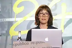 Portrait shot of a young woman behind a speaker's desk on which she has an open laptop. She is wearing glasses and looking up with a light mischievous grin. Behind her the number 24 painted in yellow is visible on the wall.