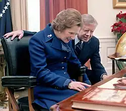 Margaret Thatcher seated behind the Resolute desk with Jimmy Carter looking over her shoulder as she read the plaque installed on the desk.