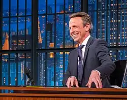 Meyers at his stage desk, in a suit, laughing