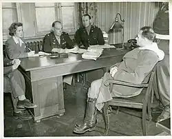 B&W photo of the US Army colonels sitting behind a wooden desk. Rudolf Hess sits in a chair obliquely facing them, right leg crossed over left, fingers interlaced in his lap.