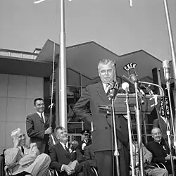 A man in suit and tie stands with arms on hips behind a podium with media microphones attached.