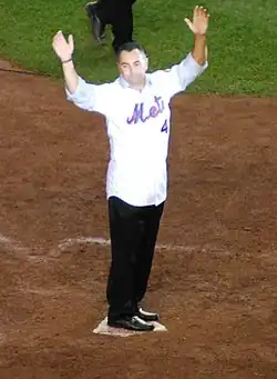 A man in a white baseball jersey and blue jeans stands on home plate with his arms upraised.