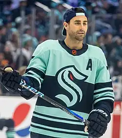 Hockey player skating while wearing a blue Seattle Kraken hockey jersey and cap while holding a stick.
